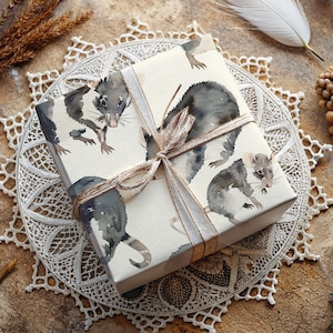 K&ouml;nnte beinhalten: Eine wei&szlig;e Geschenkbox mit einem braunen und wei&szlig;en Band. Die Box ist in Papier mit einem Aquarell-Design von schwarzen Ratten eingepackt.