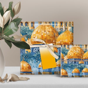 May include: Gift boxes wrapped in paper featuring a watercolor design of golden domes and blue architectural details. The boxes are tied with a cream-colored ribbon and a blank yellow tag. A white vase with green leaves is in the background.