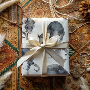 K&ouml;nnte beinhalten: Eine wei&szlig;e Geschenkbox mit einem cremefarbenen Band, das zu einer Schleife gebunden ist. Die Box ist in Papier mit einer Aquarell-Illustration von zwei schwarz-wei&szlig;en Opossums verpackt.