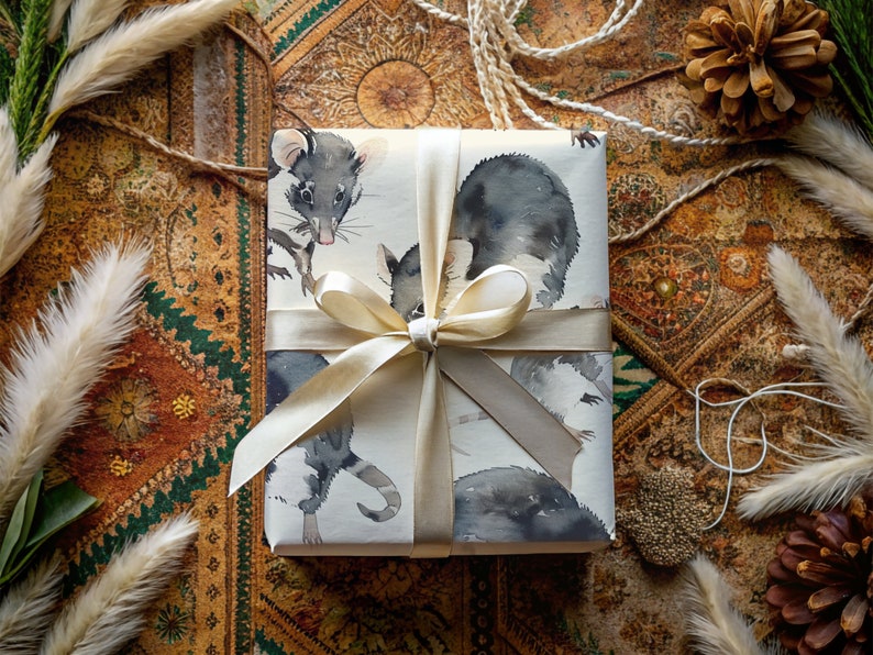 K&ouml;nnte beinhalten: Eine wei&szlig;e Geschenkbox mit einem cremefarbenen Band, das zu einer Schleife gebunden ist. Die Box ist in Papier mit einer Aquarell-Illustration von zwei schwarz-wei&szlig;en Opossums verpackt.
