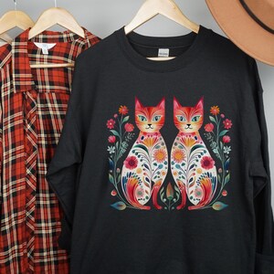 May include: Black sweatshirt with a colorful graphic of two cats with floral patterns. The cats are facing forward and have orange fur with green eyes.