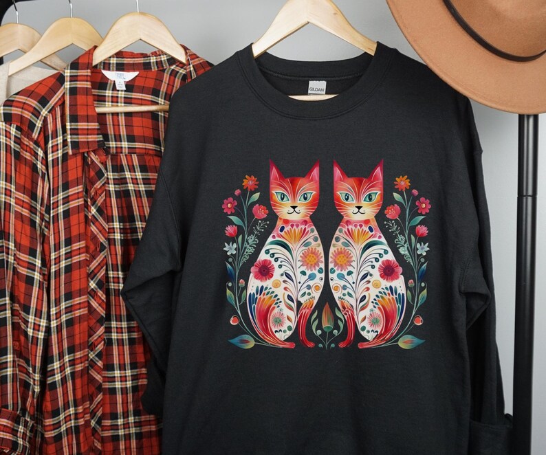 May include: Black sweatshirt with a colorful graphic of two cats with floral patterns. The cats are facing forward and have orange fur with green eyes.