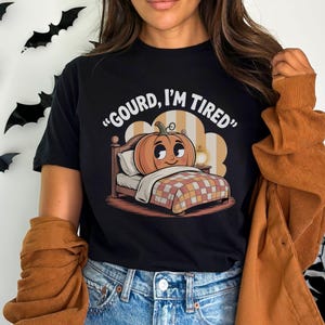 Puede incluir: Camiseta negra con un diseño de calabaza en una cama y el texto "GOURD, I'M TIRED". La calabaza tiene una cara sonriente y descansa sobre una cama con una manta a cuadros. Una prenda informal.