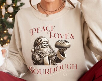 May include: Beige sweatshirt with a graphic of Santa Claus holding a loaf of bread. The text "PEACE LOVE & SOURDOUGH" is printed in burgundy script. The sweatshirt is a neutral color, suitable for the holiday season.