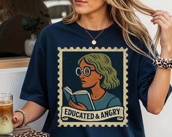 Op de afbeelding: Marineblauw T-shirt met een postzegelontwerp. De postzegel toont een vrouw met een bril, groen haar en een boek, met de tekst "EDUCATED & ANGRY". Het T-shirt heeft een casual stijl.