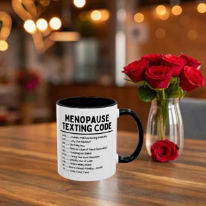 May include: A white ceramic mug with a black interior and handle, featuring the text "MENOPAUSE TEXTING CODE" and a list of acronyms and their meanings. The mug is on a wooden table, with a vase of red roses in the background.