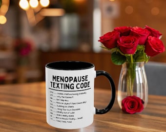 Puede incluir: Taza de cerámica blanca con interior y asa negros, con el texto "MENOPAUSE TEXTING CODE" y una lista de acrónimos. La taza está sobre una mesa de madera, con un jarrón de rosas rojas al fondo.