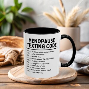 May include: A white ceramic mug with a black interior and handle. The mug features the text "MENOPAUSE TEXTING CODE" in black, along with a list of acronyms and their meanings. The mug is on a wooden tray.