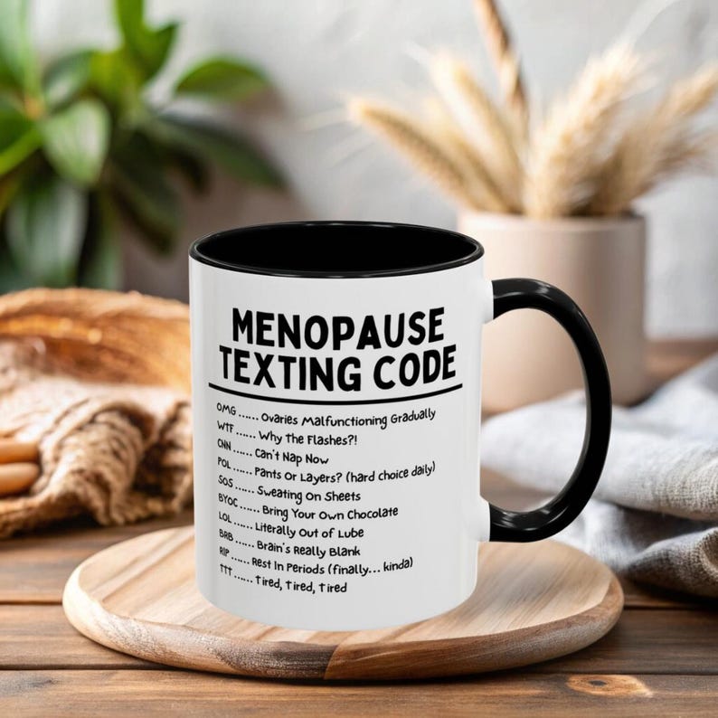 May include: A white ceramic mug with a black interior and handle. The mug features the text "MENOPAUSE TEXTING CODE" in black, along with a list of acronyms and their meanings. The mug is on a wooden tray.