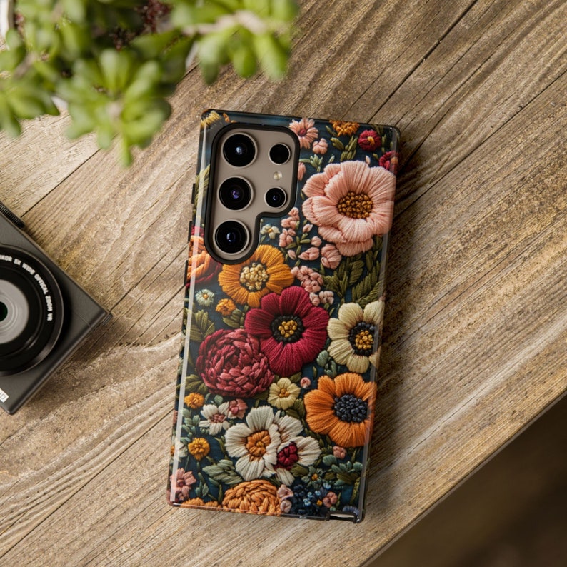 Puede incluir: Una funda para tel&eacute;fono con un dise&ntilde;o floral. La funda es negra con una variedad de flores coloridas bordadas en ella. La funda del tel&eacute;fono tiene un recorte para el objetivo de la c&aacute;mara con un borde plateado.