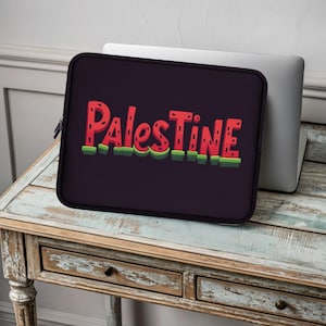 Könnte beinhalten: Eine schwarze Laptoptasche mit dem Wort "Palestine" in roter, wassermelonenartiger Schrift. Die Tasche hat einen Reißverschluss und liegt auf einem Holztisch mit einem Laptop dahinter. Der Text hat einen grünen Rand.