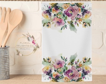 May include: A white kitchen towel with a watercolour floral design. The flowers are in shades of purple, pink, yellow, and green. The towel has a textured surface and is hanging from a rope.