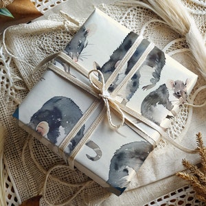 K&ouml;nnte beinhalten: Eine wei&szlig;e Geschenkbox mit braunem Band und einem Aquarell-Design aus schwarzen und wei&szlig;en Opossums. Die Box ist in ein wei&szlig;es Spitzengewebe eingewickelt.