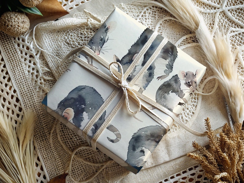 K&ouml;nnte beinhalten: Eine wei&szlig;e Geschenkbox mit braunem Band und einem Aquarell-Design aus schwarzen und wei&szlig;en Opossums. Die Box ist in ein wei&szlig;es Spitzengewebe eingewickelt.