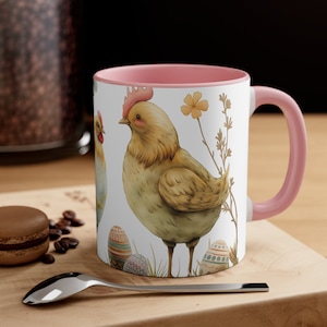 Puede incluir: Una taza de cerámica rosa y blanca con un diseño de dos gallinas y huevos de Pascua. Las gallinas son marrones y amarillas sobre un fondo blanco. Los huevos están decorados con patrones coloridos.