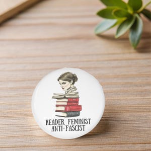 Może przedstawiać: Biały guzik z czarno-białym obrazkiem kobiety czytającej stos książek. Tekst "Reader, Feminist Anti-Fascist" jest nadrukowany czarnym kolorem pod obrazkiem.