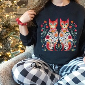 May include: Black sweatshirt with a colorful floral design featuring two cats. The cats are facing each other and have intricate patterns on their bodies. The design is surrounded by flowers and leaves.