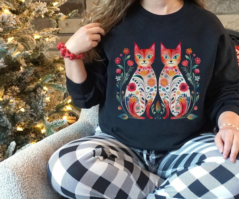May include: Black sweatshirt with a colorful floral design featuring two cats. The cats are facing each other and have intricate patterns on their bodies. The design is surrounded by flowers and leaves.