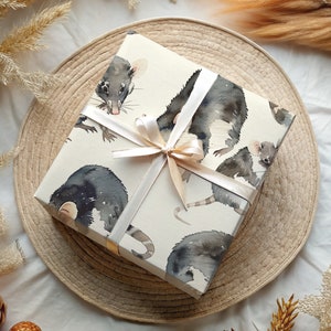 K&ouml;nnte beinhalten: Eine wei&szlig;e Geschenkbox mit braunem Band und einem Muster aus Aquarell-Illustrationen von schwarzen Ratten. Die Box liegt auf einem geflochtenen Stroh-Untersetzer.