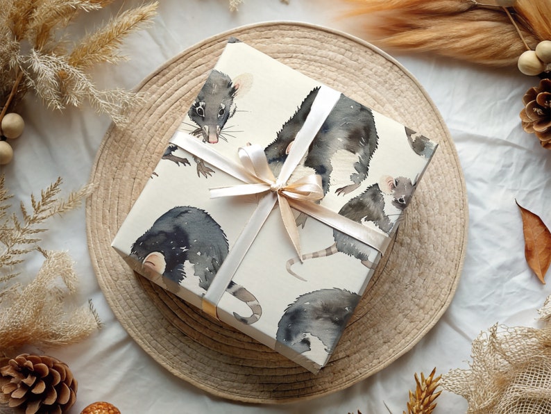 K&ouml;nnte beinhalten: Eine wei&szlig;e Geschenkbox mit braunem Band und einem Muster aus Aquarell-Illustrationen von schwarzen Ratten. Die Box liegt auf einem geflochtenen Stroh-Untersetzer.