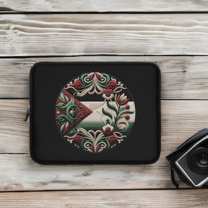 Puede incluir: Funda para portátil negra con un diseño circular que presenta un patrón floral estilizado en tonos verdes, rojos y blancos. El diseño se asemeja a una bandera con una forma triangular.