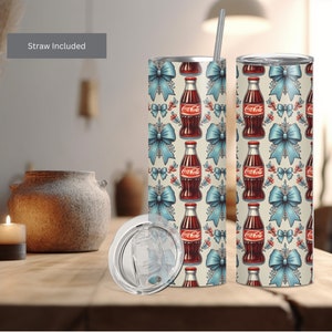 Könnte beinhalten: Ein Edelstahlbecher mit weißem Hintergrund und einem Muster aus blauen Schleifen, roten Stechpalmenbeeren und Coca-Cola-Flaschen. Der Becher hat einen durchsichtigen Deckel und einen Strohhalm. "Straw Included" steht oben im Bild.
