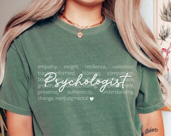 May include: Sage green t-shirt with the word "Psychologist" in a cursive font, surrounded by related words like "empathy" and "compassion." The shirt has a crew neck and short sleeves.