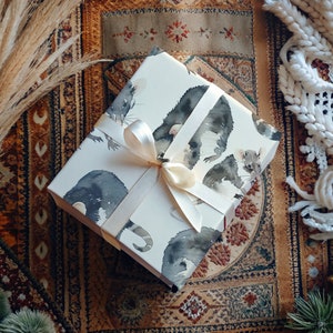 K&ouml;nnte beinhalten: Wei&szlig;e Geschenkpapier mit einem Aquarell-Design aus schwarzen und wei&szlig;en M&auml;usen. Das Geschenkpapier ist mit einem wei&szlig;en Satinband gebunden.