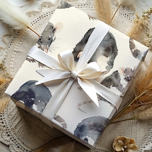 K&ouml;nnte beinhalten: Wei&szlig;e Geschenkpapier mit einem Aquarell-Design von schwarzen und grauen M&auml;usen. Das Geschenkpapier ist mit einem wei&szlig;en Satinband gebunden.