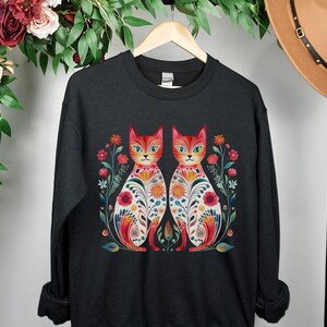 May include: Black sweatshirt with a colorful illustration of two cats with floral patterns. The cats are facing each other and have red, orange, yellow, green, and blue colors.