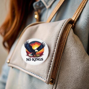 Könnte beinhalten: Ein weißer Knopf mit einem bunten Bild eines Adlers mit einer Krone in seinen Krallen vor einem Sonnenuntergangs-Hintergrund. Der Text "NO KINGS" ist unter dem Bild gedruckt.