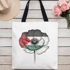 May include: A white tote bag with black handles features a black and white line drawing of a poppy flower. The flower's petals are red, green, and white, resembling a flag.