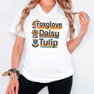 Può includere: T-shirt bianca con scollo a V con le parole "Foxglove", "Daisy" e "Tulip" in un font retrò. Ogni parola è accompagnata da un'illustrazione floreale. Il testo è in un design colorato a strati.