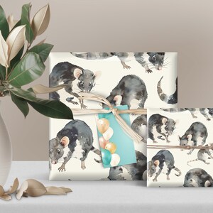 K&ouml;nnte beinhalten: Zwei Geschenkboxen, die in wei&szlig;es Papier mit einem sich wiederholenden Muster aus Aquarell-Illustrationen von schwarzen und wei&szlig;en M&auml;usen verpackt sind. Die gr&ouml;&szlig;ere Box ist mit einem braunen Band gebunden und hat ein blaues Etikett mit wei&szlig;en Luftballons. Die kleinere Box ist mit einer braunen Schnur gebunden.