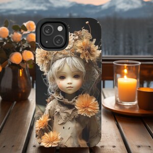 Puede incluir: Una funda de teléfono con un diseño caprichoso que presenta una muñeca con una corona de flores y un fondo rústico. La muñeca tiene el pelo rubio y los ojos azules y lleva un vestido marrón.