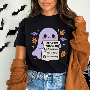 May include: Black t-shirt featuring a cute purple ghost holding a "Self-Care Checklist." The checklist includes "Drank water," "Went to therapy," and "Set a boundary." The design includes orange leaves and purple bats.