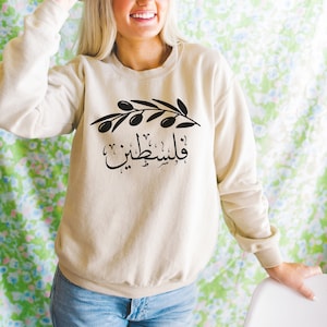 May include: A beige sweatshirt with a black olive branch and Arabic text that reads "Palestine" printed on the front.