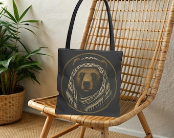 May include: Black tote bag featuring a brown bear wearing a traditional keffiyeh headdress. The bear's portrait is framed in a gold circle. The bag has black handles and is displayed on a wicker chair.