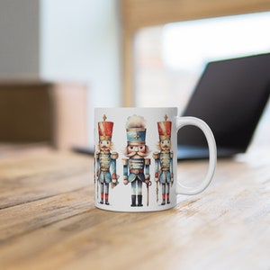 Może przedstawiać: Biały kubek ceramiczny z kolorową ilustracją trzech żołnierzyków do łamania orzechów. Żołnierzyki są ubrani w niebieskie i czerwone mundury z złotymi akcentami.