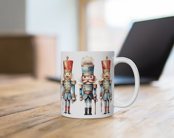 May include: White ceramic mug with a colorful illustration of three nutcracker soldiers. The nutcrackers are wearing blue and red uniforms with gold accents.