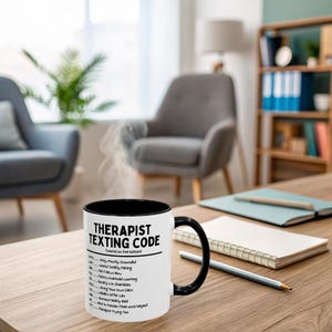 May include: A white ceramic mug with a black interior and handle. The mug features the text "THERAPIST TEXTING CODE" and a list of phrases. The mug is on a wooden table with a notebook and pencil.