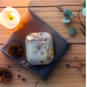 Op de afbeelding: Een witte Airpods-hoes met een ontwerp van geperste bloemen. De hoes heeft gedroogde bloemen in tinten van bruin, blauw en wit.