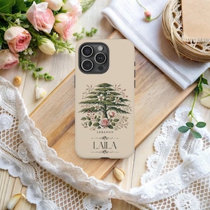 May include: Beige phone case featuring a detailed illustration of a cedar tree with flowers and the word "LEBANON". The name "LAILA" is printed below the tree. The case is surrounded by flowers and lace.