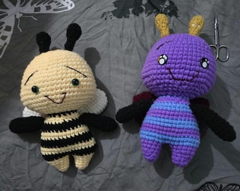 May include: Two handmade crochet insect plush toys. One is a bee with a cream-colored head and black and cream striped body. The other is a purple insect with blue stripes. Both have black wings and smiling faces. A pair of scissors is in the background.