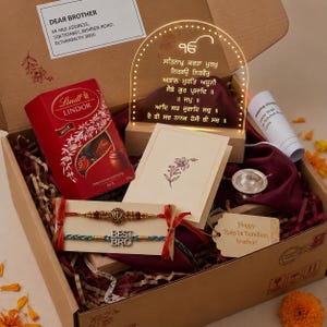 May include: A gift box containing a red Lindt Lindor chocolate box, an arched acrylic light with Gurmukhi script, a greeting card with a floral sketch, two Rakhi bracelets with 'BEST BRO' text, a small silver bowl, and a gift tag reading 'Happy Raksha Bandhan, brother!'
