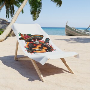 May include: A white beach towel with a colorful illustration of a skeleton playing a guitar. The skeleton is wearing a sombrero and has flowers in its hair. The towel is on a wooden beach chair.