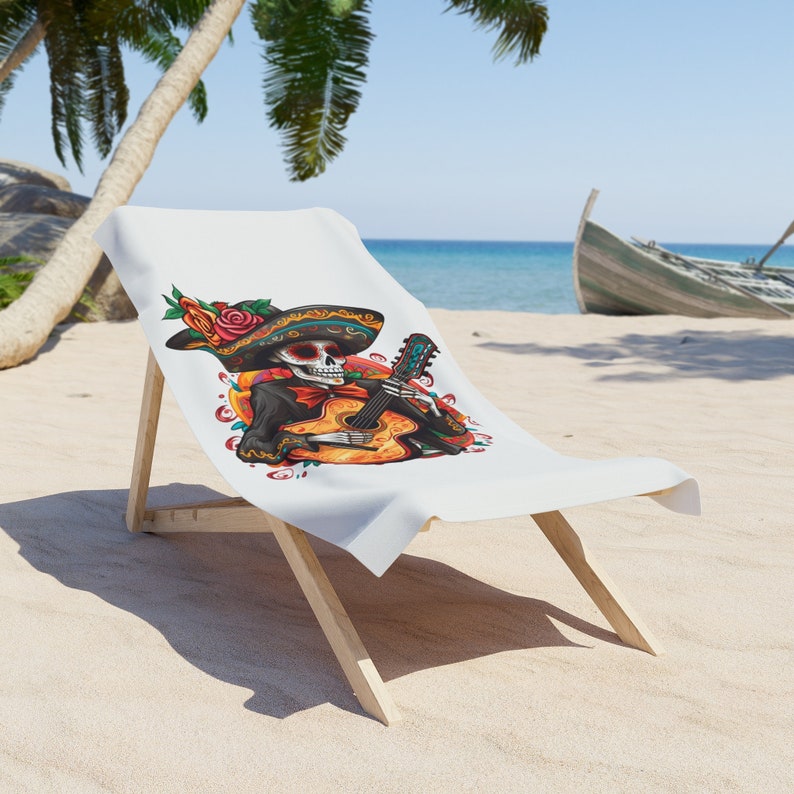 May include: A white beach towel with a colorful illustration of a skeleton playing a guitar. The skeleton is wearing a sombrero and has flowers in its hair. The towel is on a wooden beach chair.