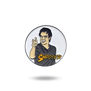 Puede incluir: Un pin de esmalte redondo con una ilustración de dibujos animados de un hombre con cabello oscuro y una camisa negra. El hombre está apuntando con su mano derecha y sonriendo. El texto "SHOOTER" está escrito en letras amarillas y naranjas debajo del hombre.