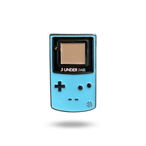May include: A blue and black enamel pin shaped like a handheld video game console. The console has a screen with a brown background and the text "3 UNDER PAR" on the front.