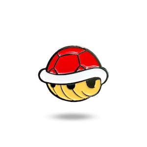 May include: A red and yellow enamel pin shaped like a Super Mario Bros. Koopa shell. The shell is closed and has a white rim.